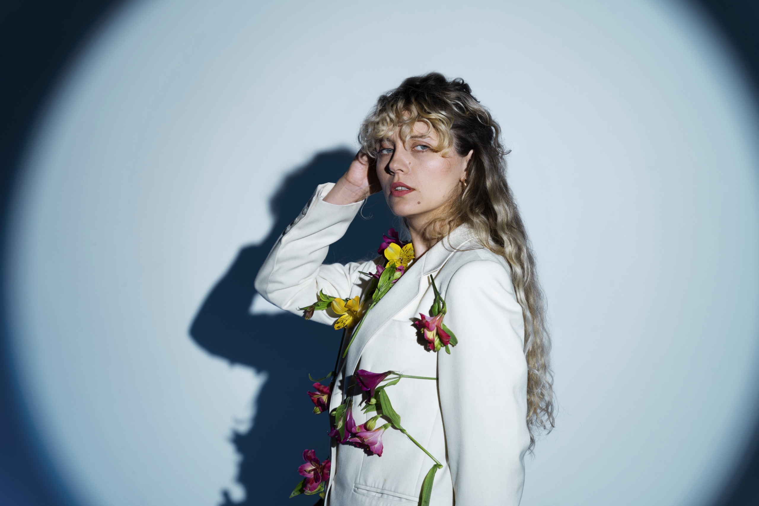 Model posing in a creative studio photo shoot wearing a white blazer decorated with flowers, under dramatic spotlight lighting.